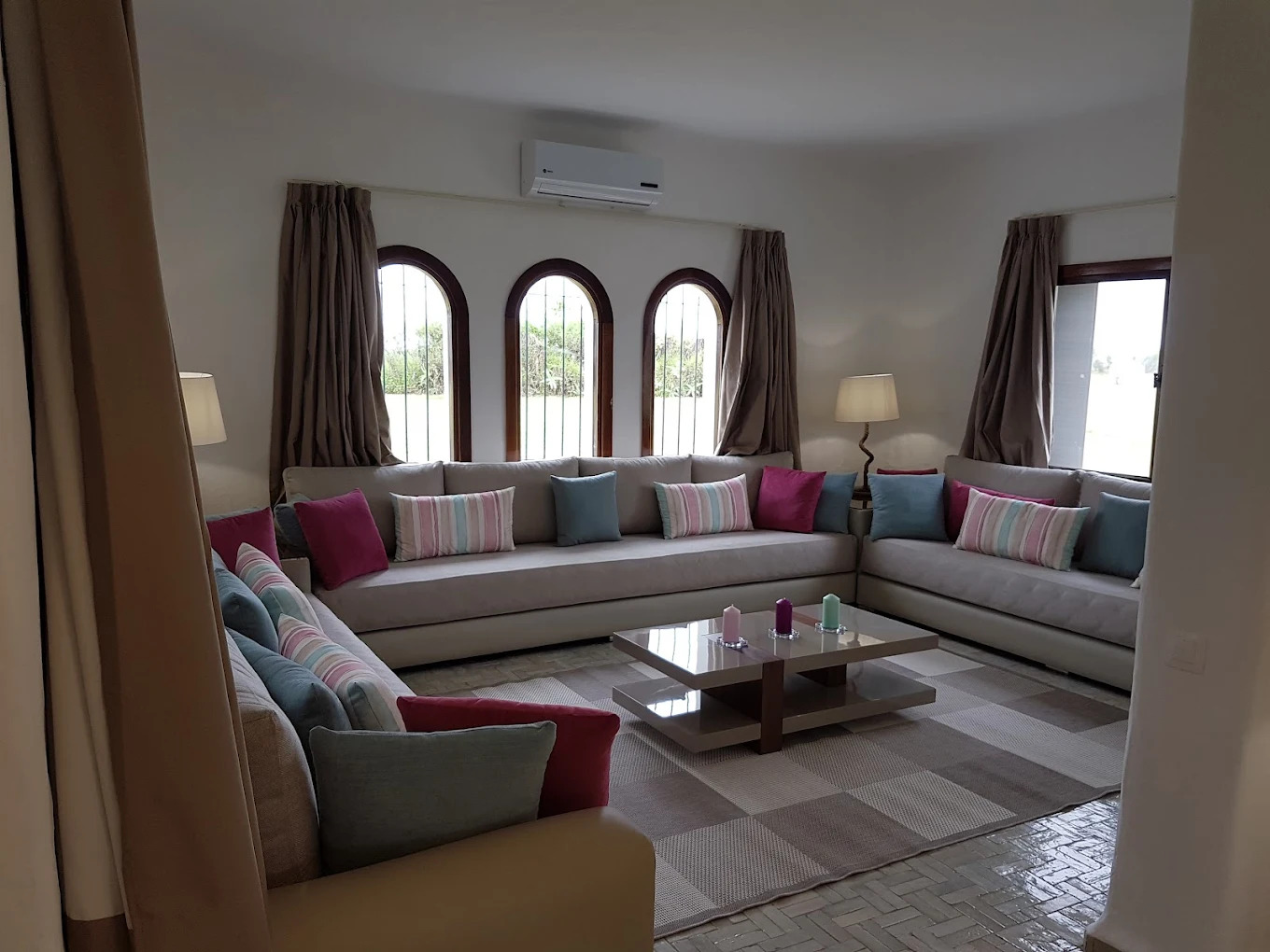 Salon marocain avec banquettes beiges, coussins colorés et fenêtres en arc